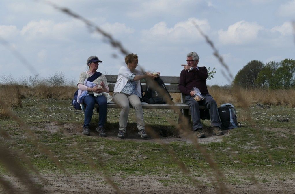 Drie mensen zitten op een bankje in een veld; twee van hen zijn aan het eten. Bewolkte lucht.