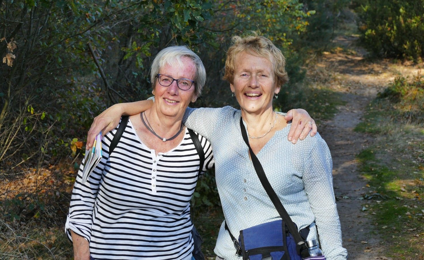 Twee vrouwen glimlachen en staan ​​met hun armen om elkaar heen op een zandpad, omringd door bomen.