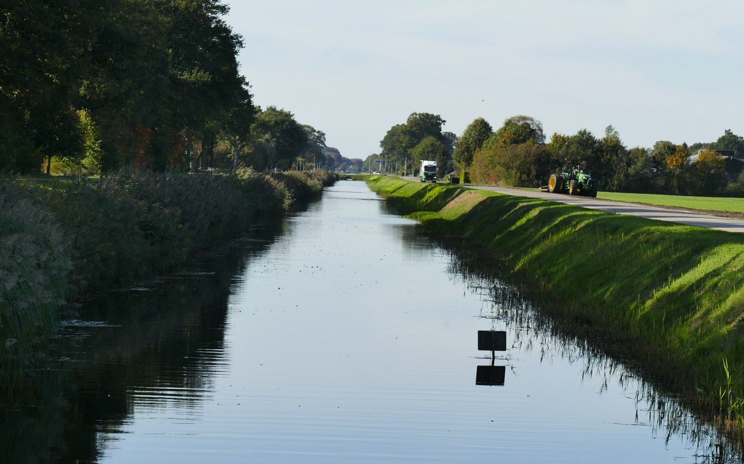 Een smal, rustig kanaal , geflankeerd door groene, met gras begroeide oevers, bomen en een weg.