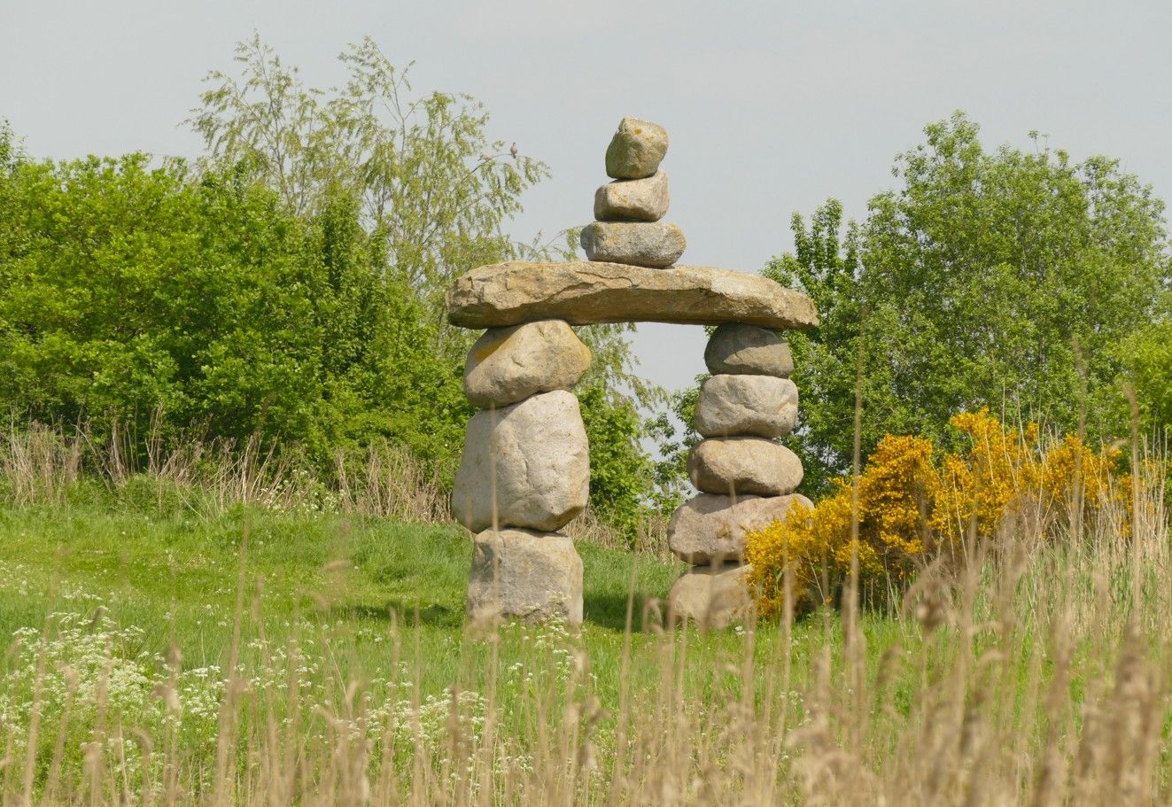 Een stenen boog, opgebouwd uit opgestapelde stenen, in een grasveld met bomen op de achtergrond.