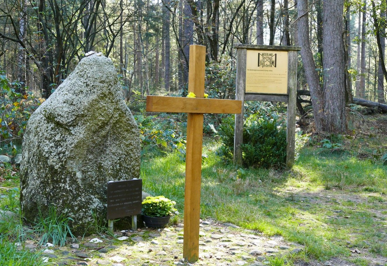 In een bos staat een houten kruis naast een grote gedenksteen en een houten informatiebord.