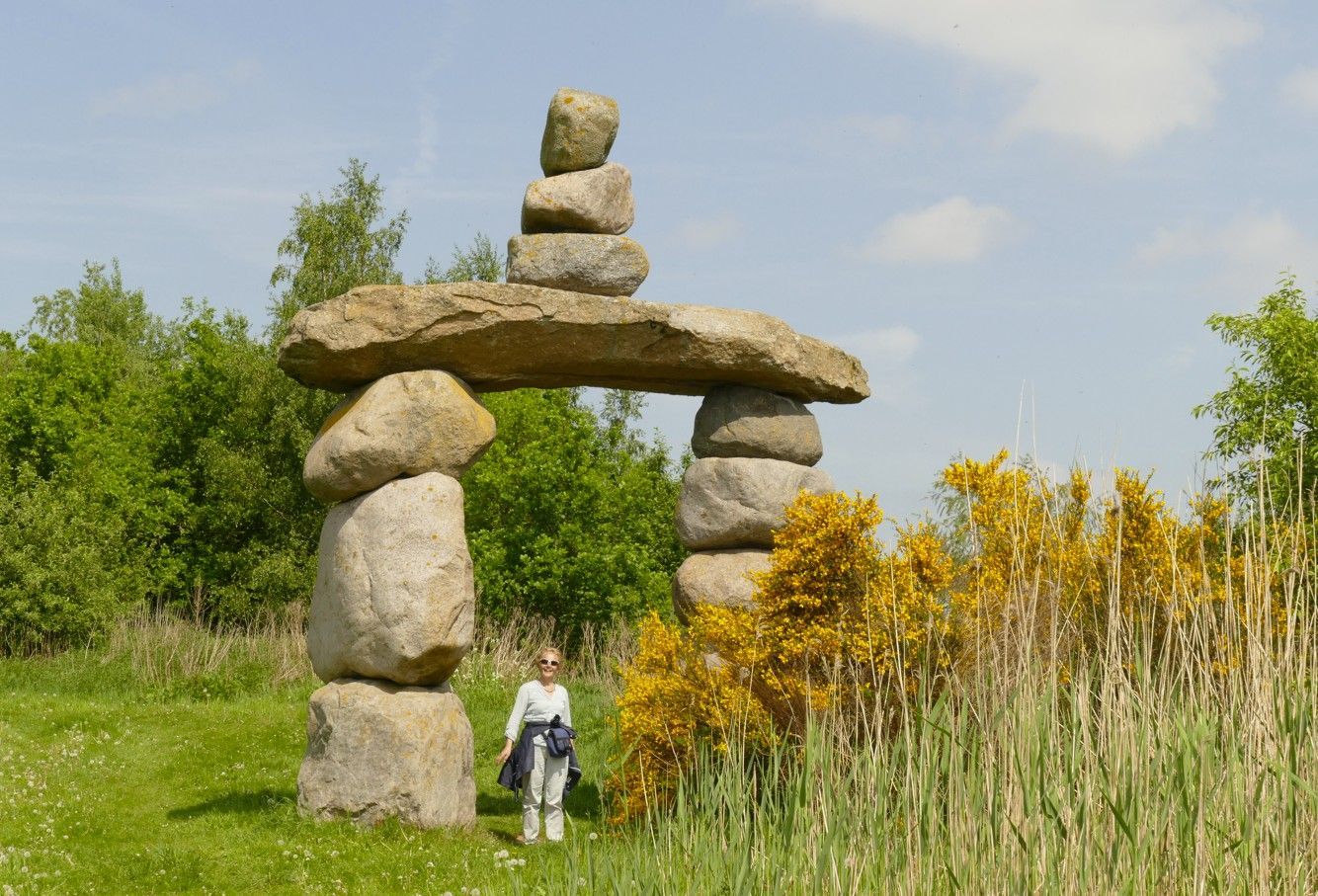 Een stenen boogsculptuur in een grasveld met een persoon eronder. Een heldere hemel, groen gebladerte en gele bloemen.