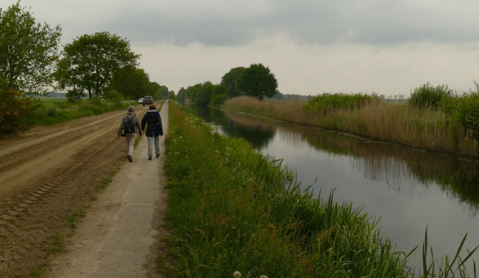 Twee mensen lopen over een zandpad langs een kanaal. Bewolkte lucht, groene rietstengels, bomen.