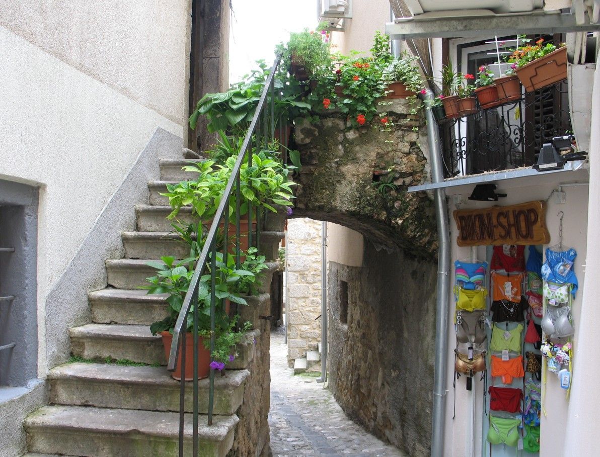 Een stenen trap leidt naar een gewelfde doorgang in een steegje. Potten met planten en een winkelbord zijn zichtbaar.
