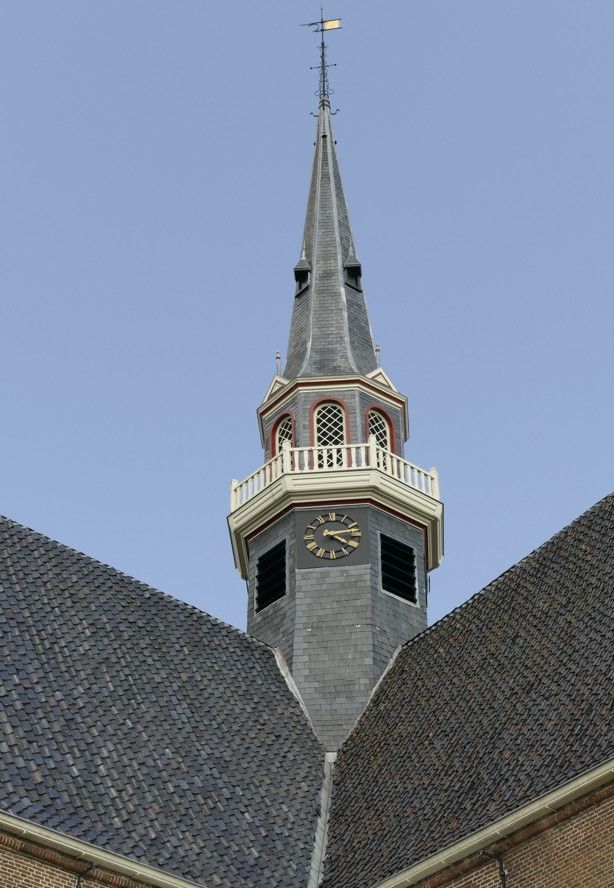 Kerktoren met klok, wit balkon en windwijzer tegen een blauwe hemel.