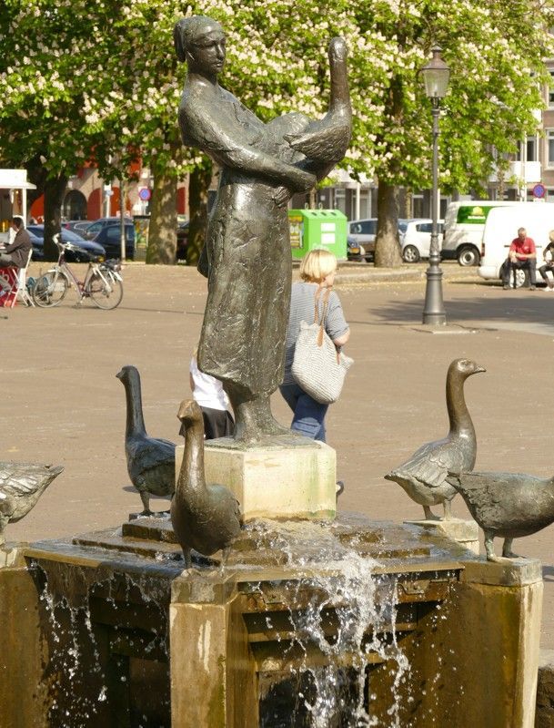 Bronzen beeld van een vrouw met een gans, omringd door ganzenbeelden in een fontein.
