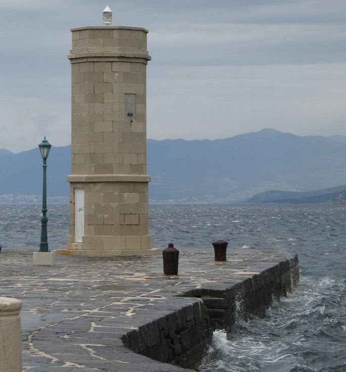 Stenen vuurtoren op een pier, met een lantaarnpaal. Ruwe zee, bergen in de verte. Bewolkte lucht.