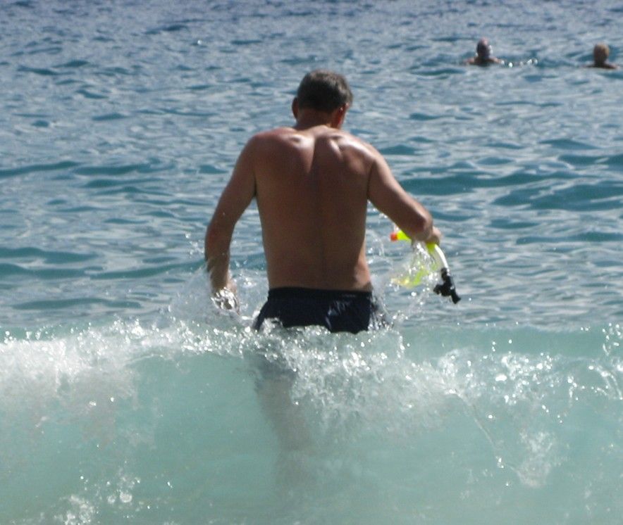 Een man waadt met snorkeluitrusting de oceaan in; helderblauw water en een zonnige dag.