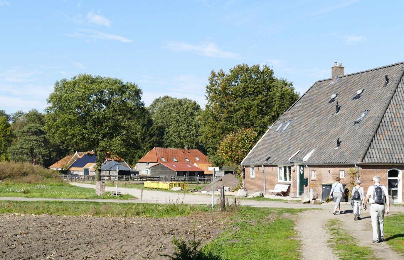 Op een zonnige, landelijke dag lopen drie mensen over een zandpad richting een bakstenen boerderij.