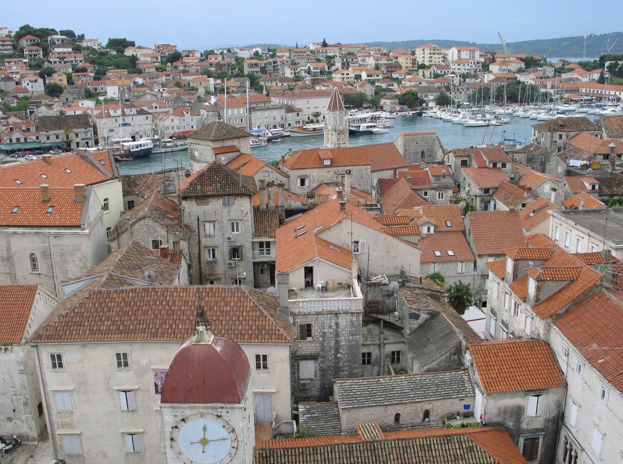 Bovenaanzicht van een Europese kuststad met rode pannendaken, haven en klokkentoren.