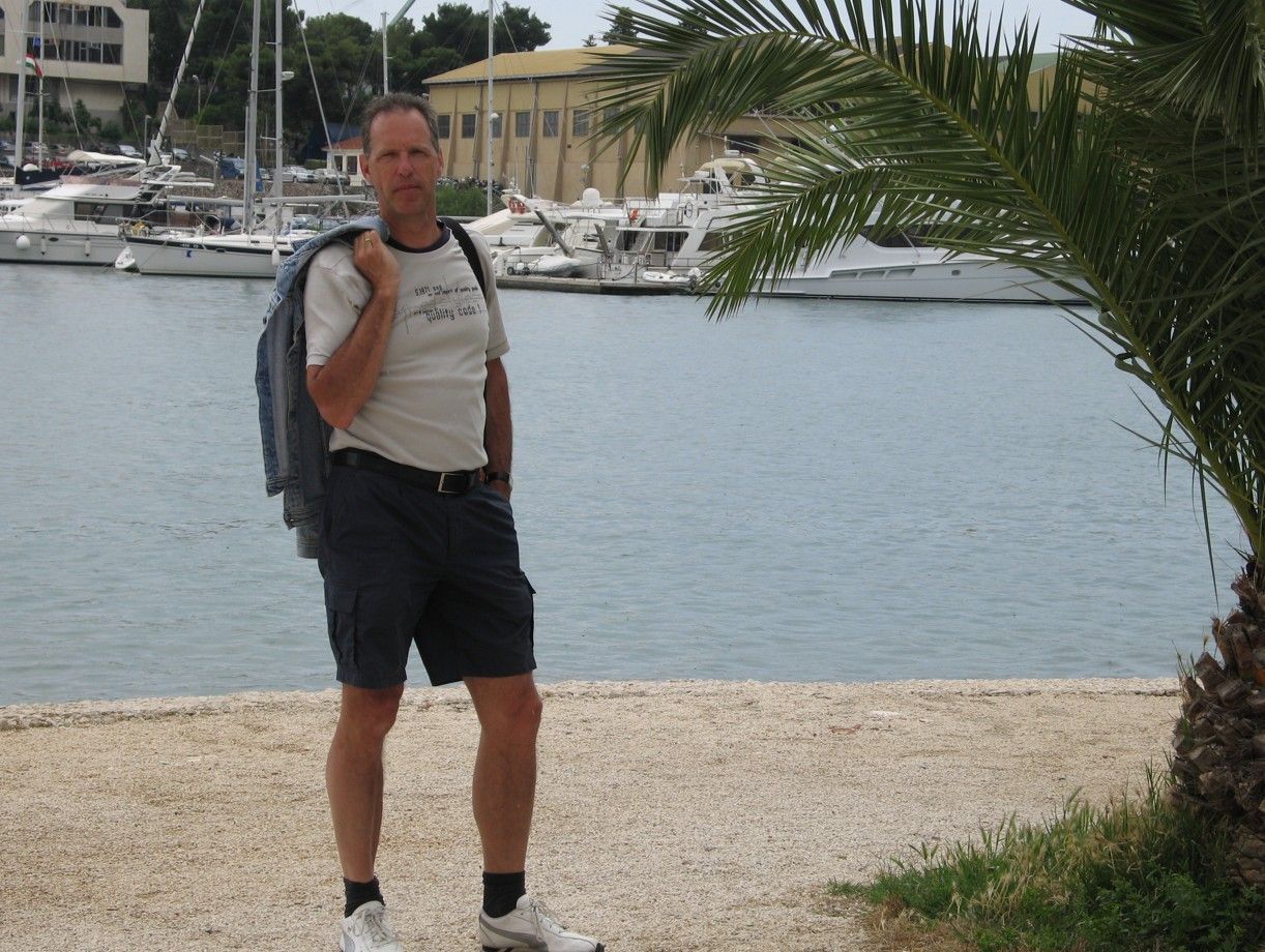 Man in korte broek staat bij het water, jas over de schouder, boten op de achtergrond.