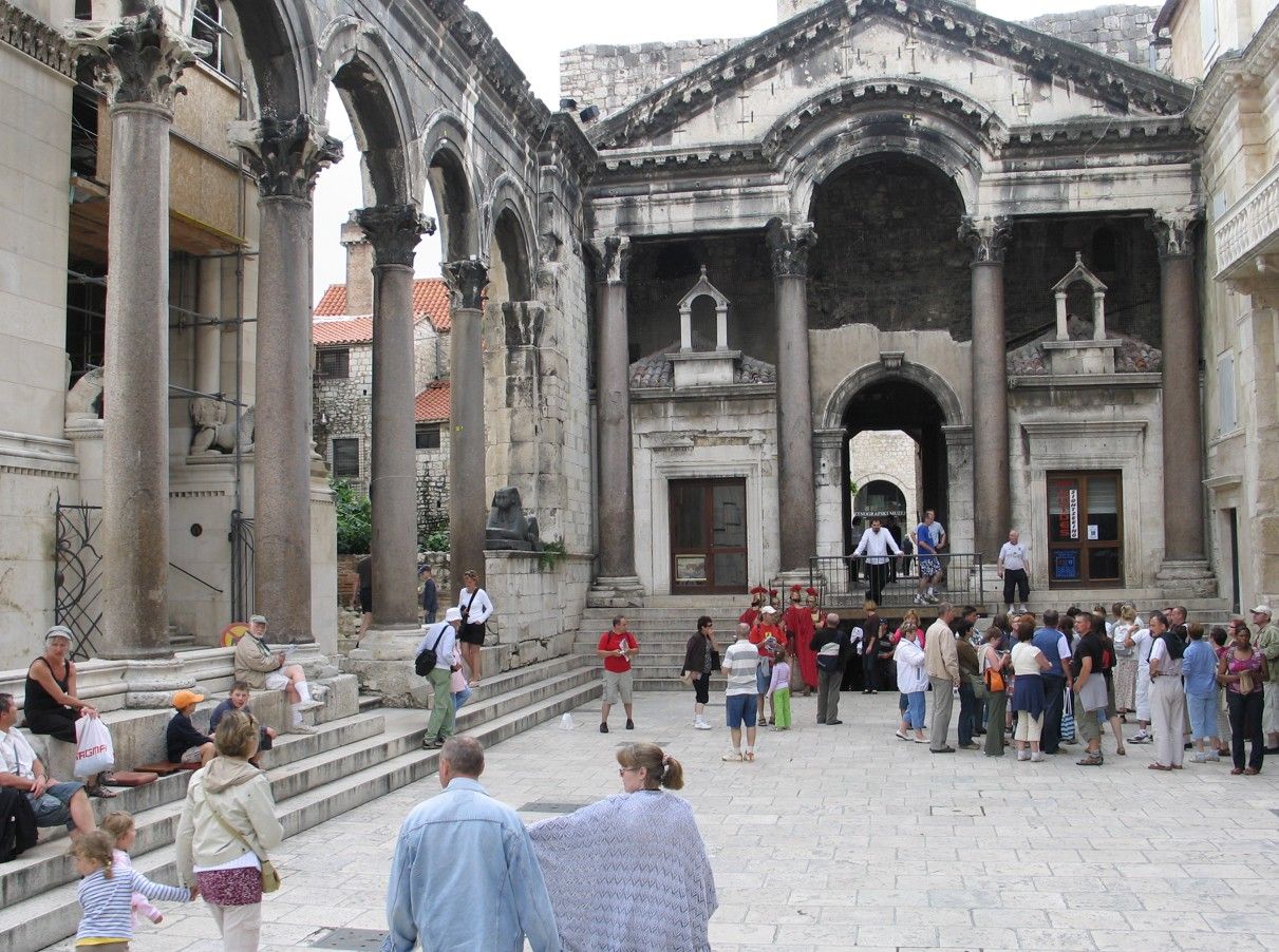 Oude stenen gevel met zuilen en bogen, vol mensen, zonnige dag.