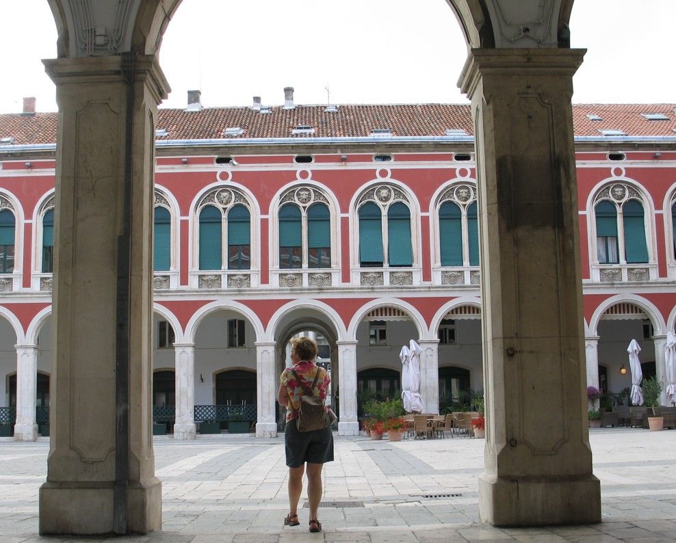 Een vrouw staat in een gewelfde doorgang en kijkt naar een rood gebouw met boogvormige ramen.