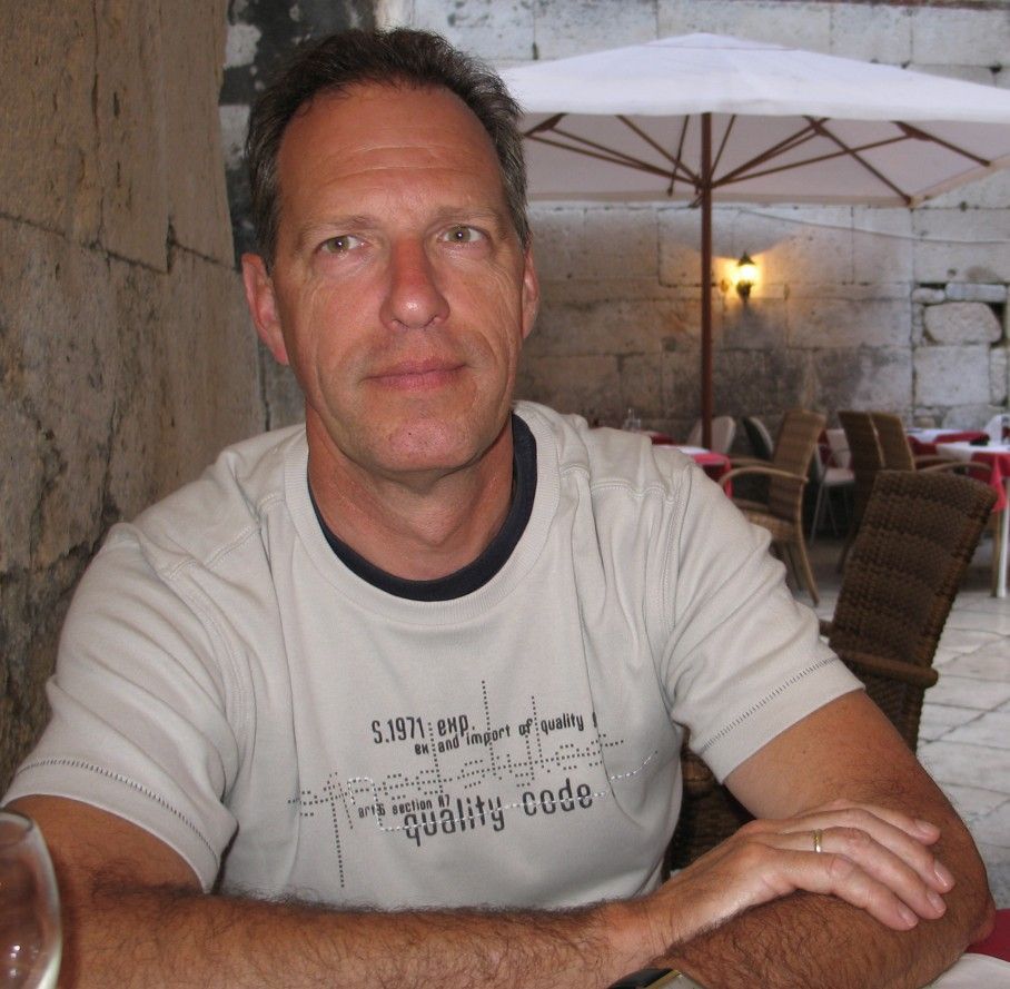 Man zit buiten aan een tafeltje, gekleed in een T-shirt; neutrale uitdrukking.