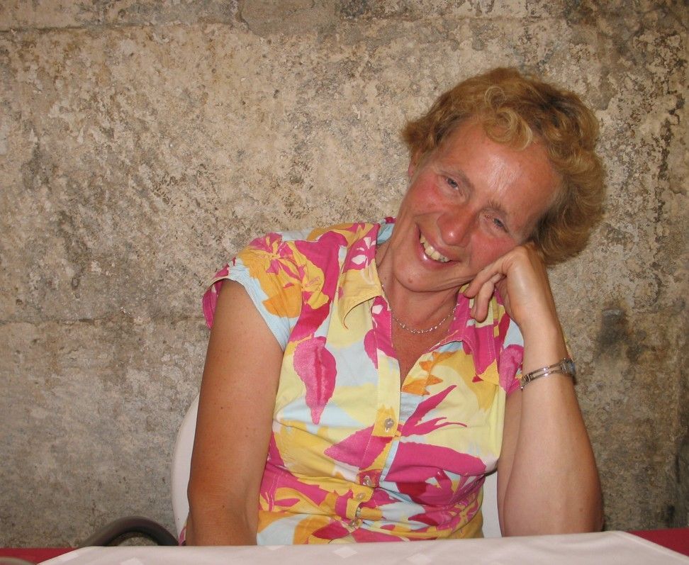 Een vrouw in een felgekleurde top met bloemenprint glimlacht en laat haar hoofd op haar hand rusten, tegen een muur van natuursteen met een ruwe structuur.