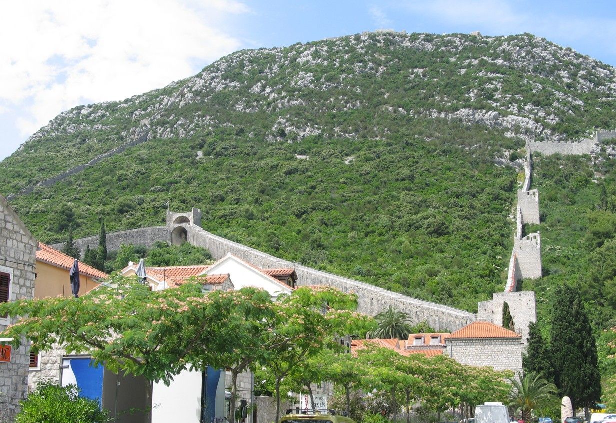Stenen muren kronkelen tegen een groene, rotsachtige heuvel op en bieden uitzicht op een dorp met rode pannendaken.