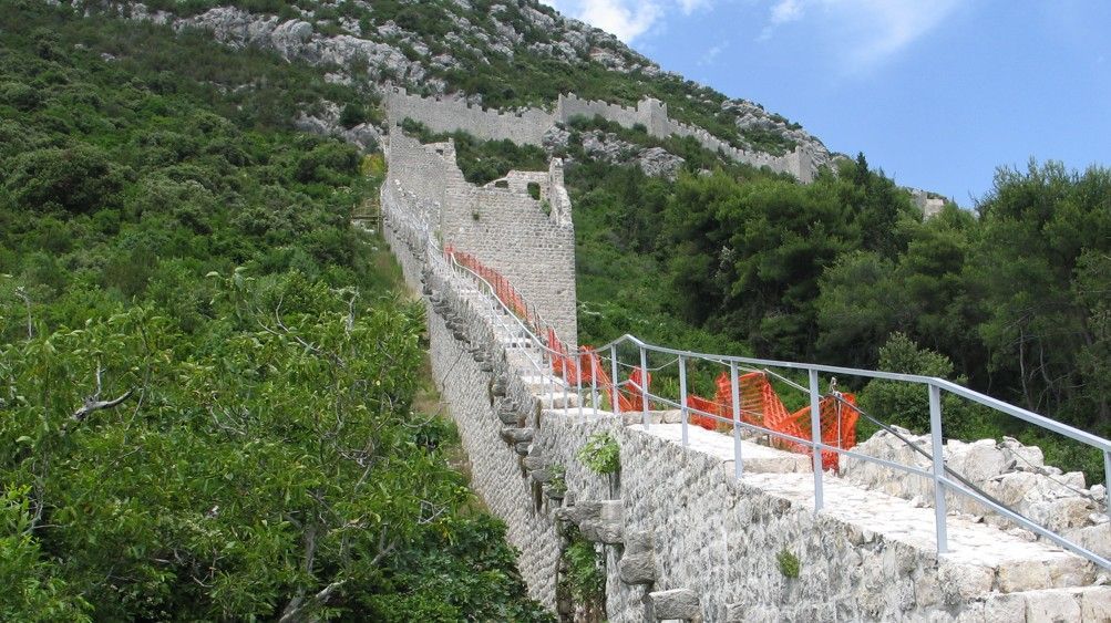 Stenen muren kronkelen tegen een berghelling op, met trappen en oranje veiligheidshekken. Omgeven door groen gebladerte.
