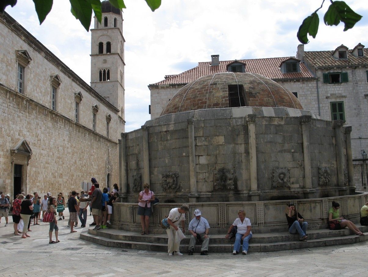De Onofrio-fontein in Dubrovnik, Kroatië, met mensenmassa's eromheen, een stenen constructie en een klokkentoren op de achtergrond.