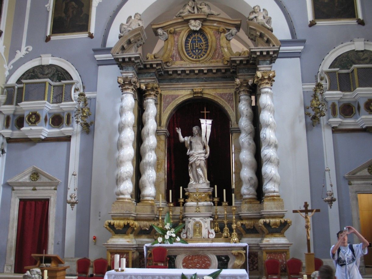 Altaar in een kerk. Een wit beeld staat in een nis, omlijst door spiraalvormige zuilen. Rode draperieën en kaarsen zijn aanwezig.
