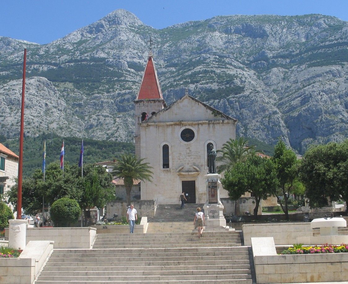 Een kerk met een rood dak en een torenspits, gelegen tegen een berg. In de buurt wapperen vlaggen, op de achtergrond staan ​​bomen.