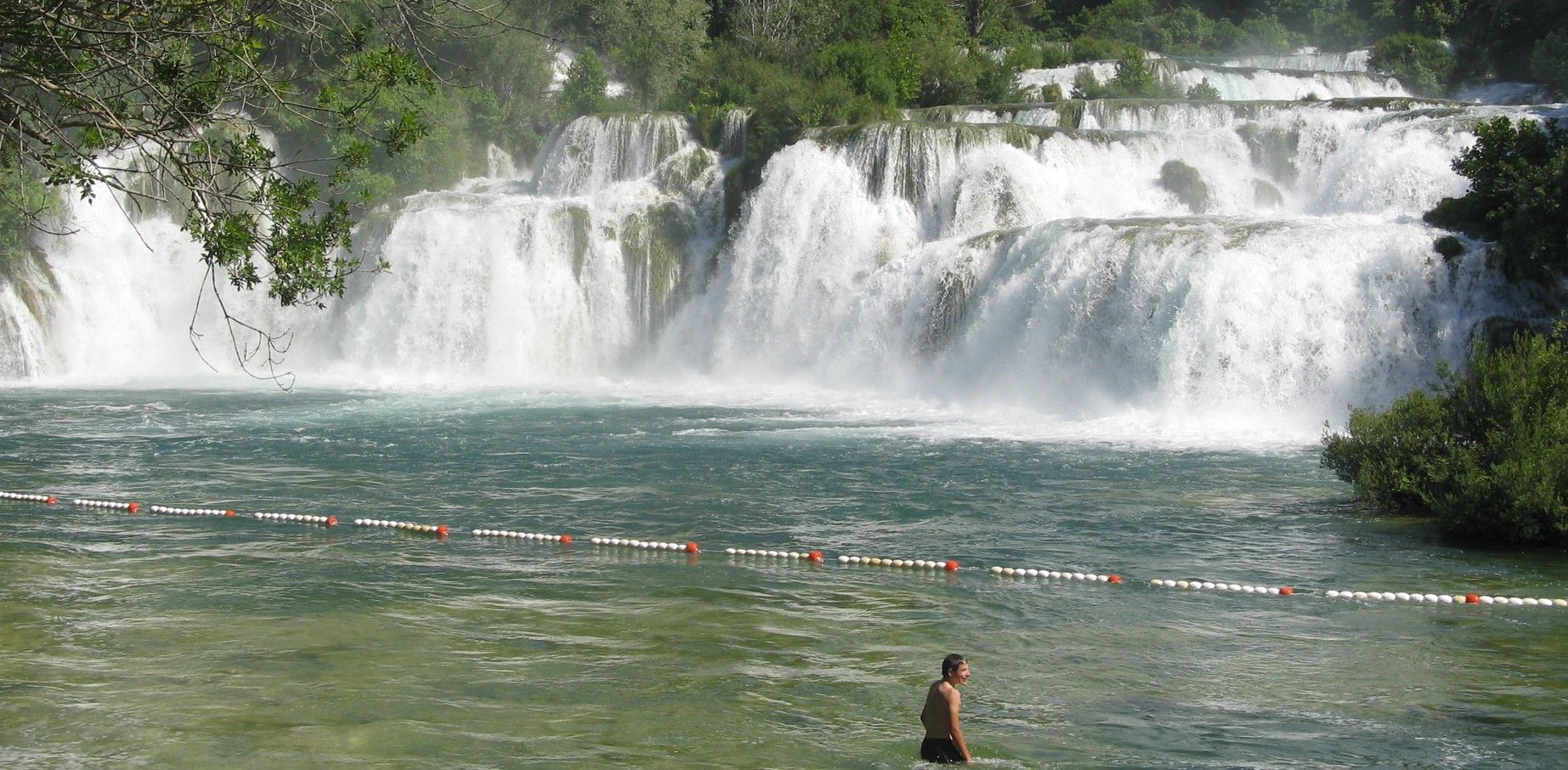 Watervallen storten neer in een waterpoel, waarin een persoon staat. Weelderig groen omringt het tafereel.