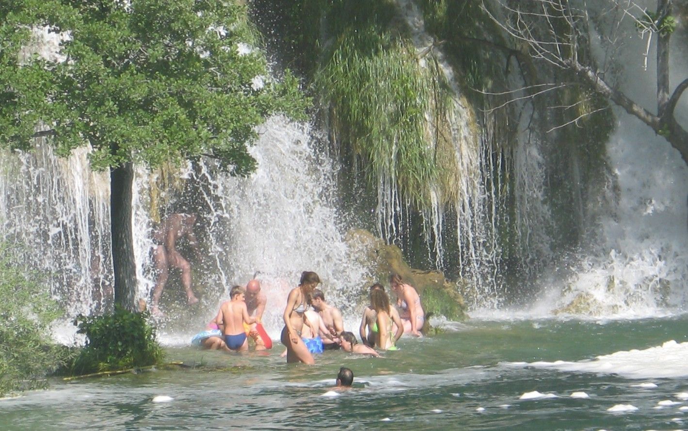 Mensen zwemmen en ontspannen onder een waterval; groen gebladerte en opspattend water; een zonnige dag.