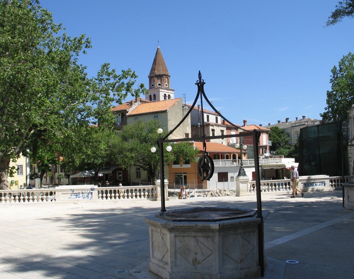 Een stenen waterput op een stadsplein met een kerktoren op de achtergrond, onder een blauwe hemel.