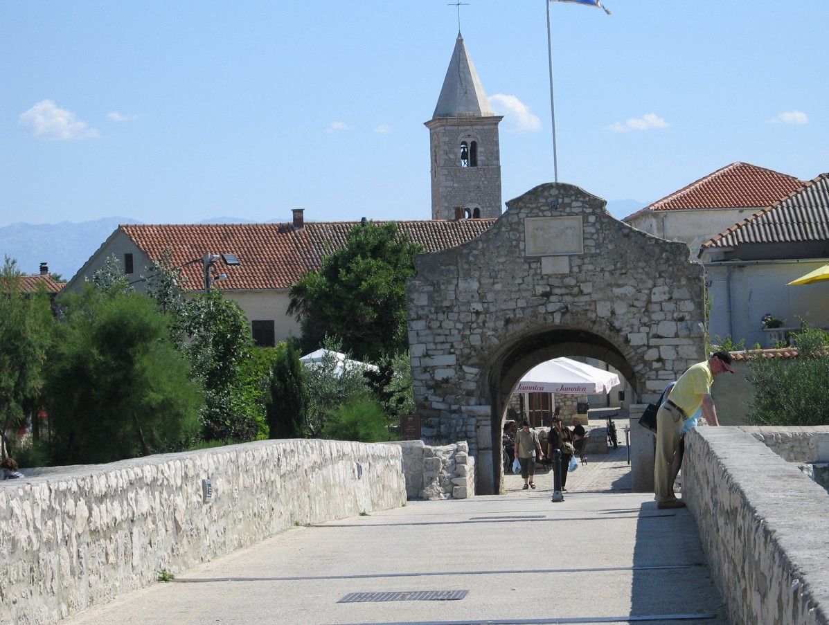 Een stenen poort die toegang geeft tot een stadsplein, met een kerktoren en mensen die erlangs lopen. Blauwe lucht.