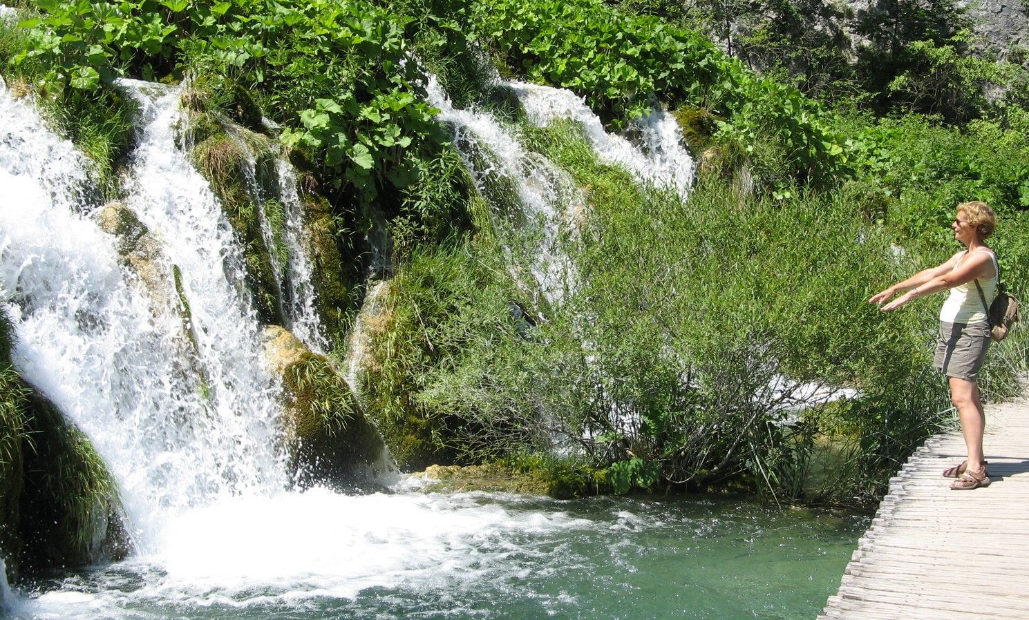 Kroatie 2008 - GOUDE OUDE Een vrouw staat bij een waterval met uitgestrekte armen. Groen gebladerte, een houten pad en turquoise water.