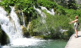 Een vrouw staat bij een waterval met uitgestrekte armen. Groen gebladerte, een houten pad en turquoise water.