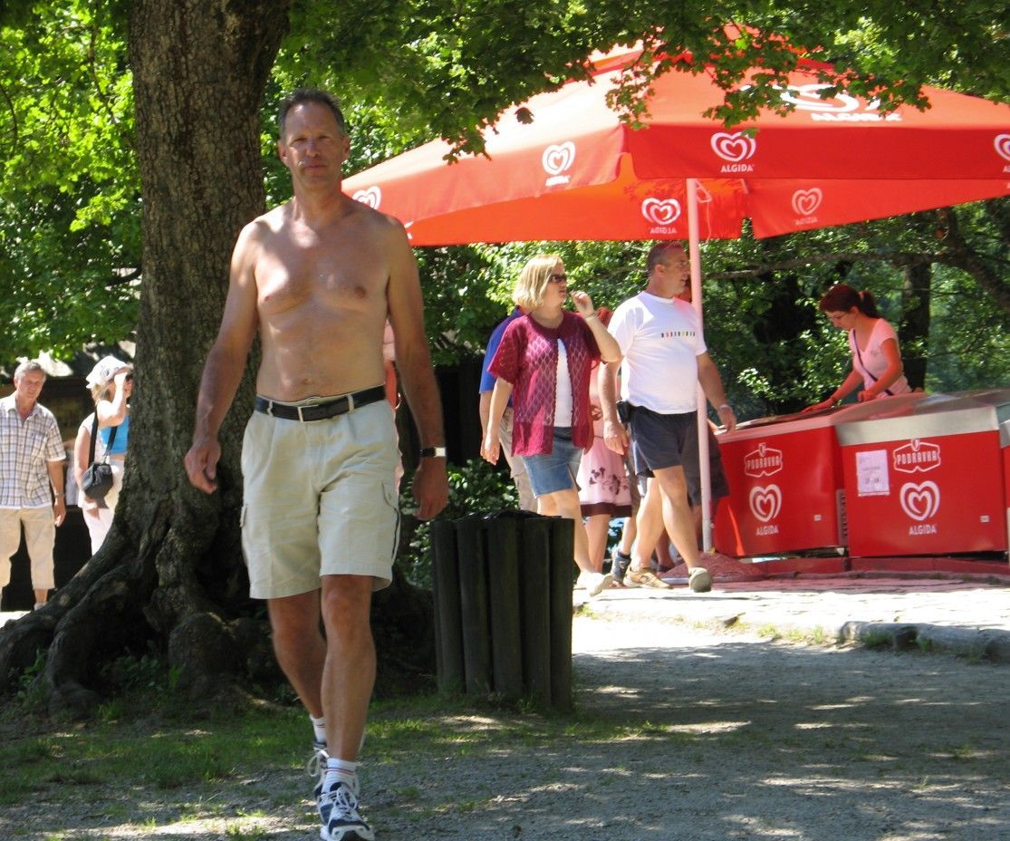 Een man in korte broek en onderhemd loopt naar de camera toe; op de achtergrond staan ​​mensen en een ijsje onder een rode paraplu.
