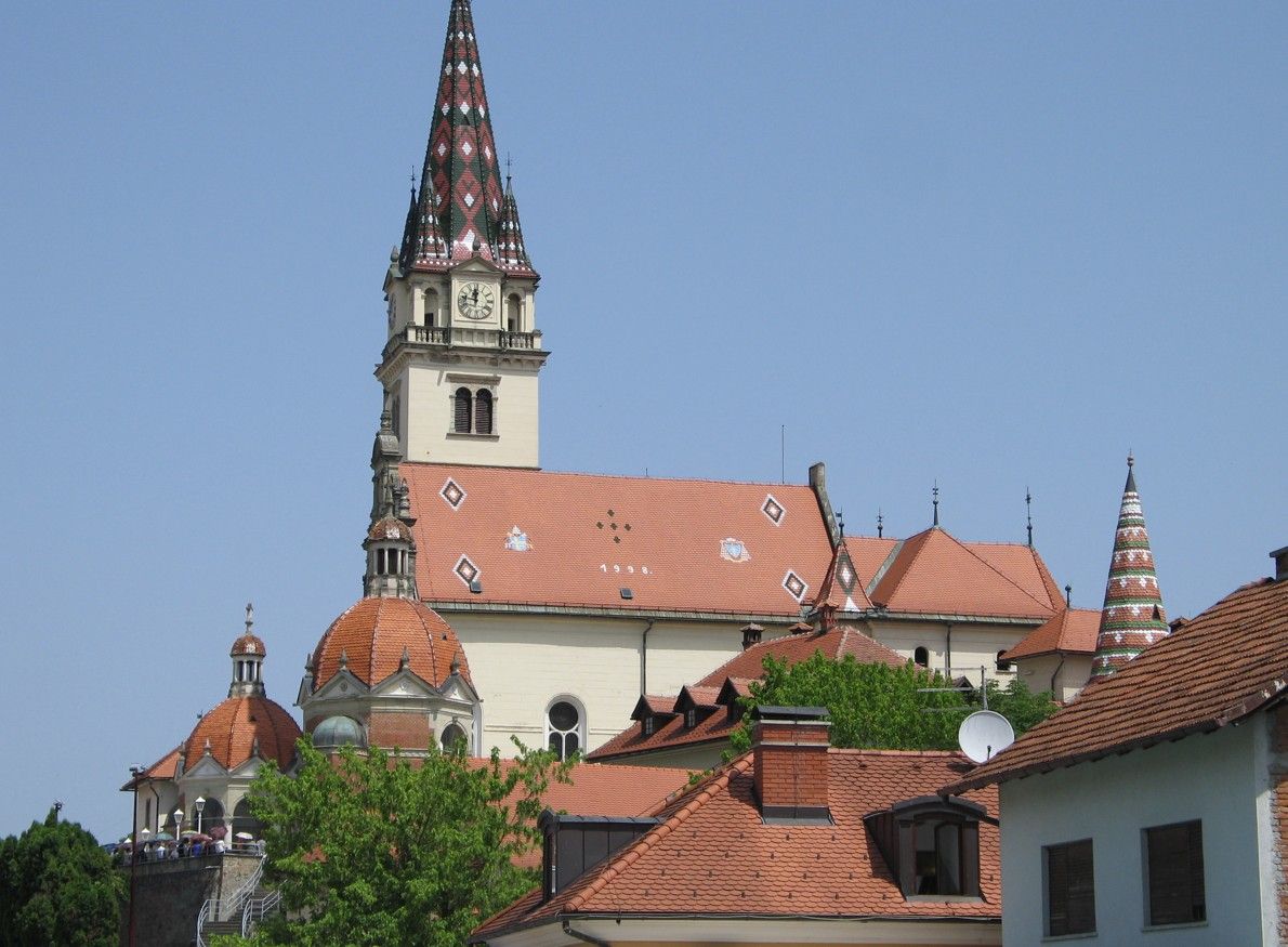 Een kerk met een rood pannendak en een hoge, sierlijke torenspits tegen een helderblauwe hemel.