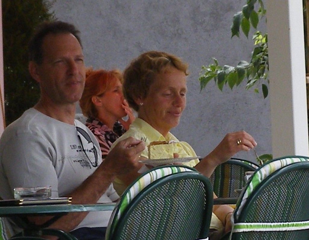Mensen zitten op een terrasje, iemand eet taart. Grijze buitenkant van het gebouw, groene stoelen.