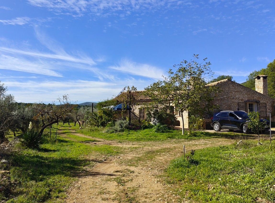 Stenen huis met een geparkeerde auto, een zandpad en bomen onder een blauwe hemel.