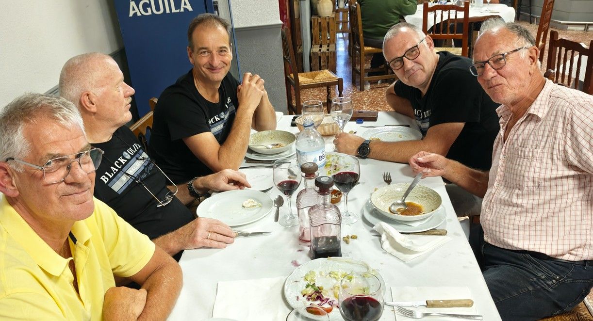 Vijf mensen zitten aan een tafel met eten en drinken en voeren een gesprek. Binnen, verlicht.