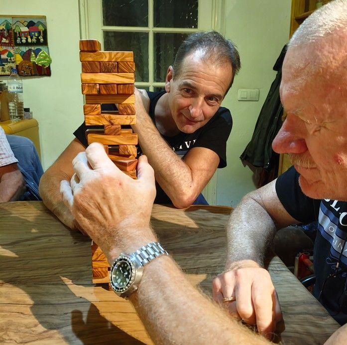 Twee mannen spelen Jenga aan een houten tafel. De ene man is geconcentreerd, de andere lacht. Binnen.