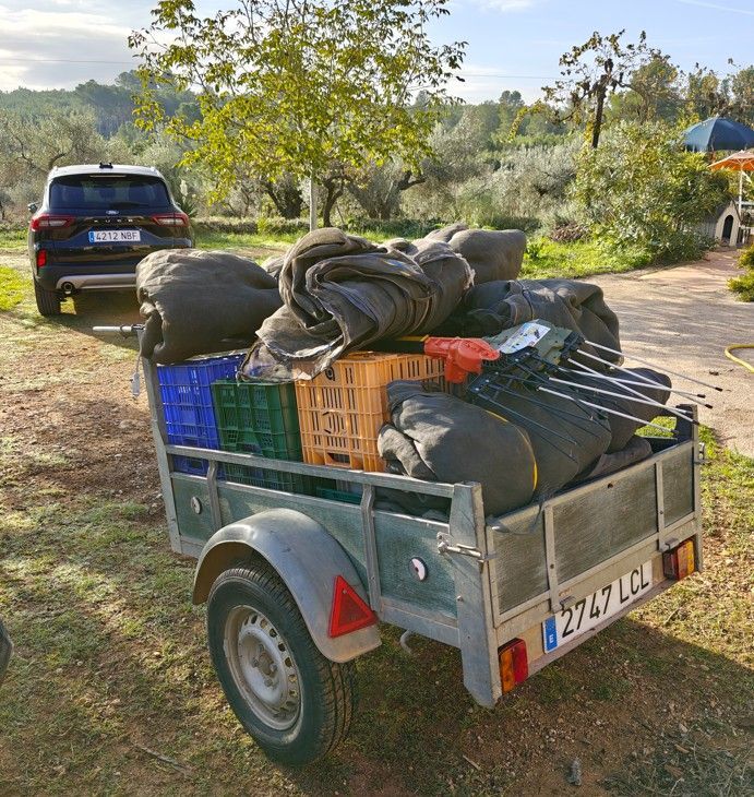 Een aanhanger volgeladen met spullen staat geparkeerd naast een auto, in een zonnige buitenomgeving met bomen en heuvels op de achtergrond.