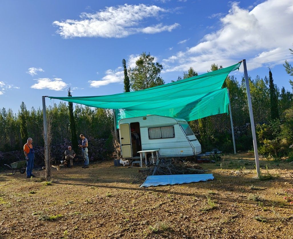 Een caravan in de schaduw van een groen bladerdak, met mensen in de buurt op een veld onder een blauwe hemel.