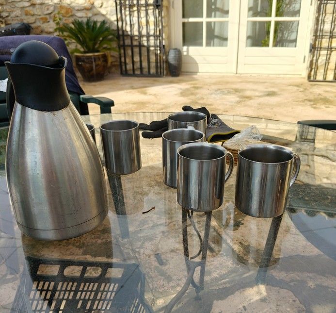 Een zilveren koffiekan en mokken staan ​​op een glazen tafel buiten bij een deur, met handschoenen op de achtergrond.
