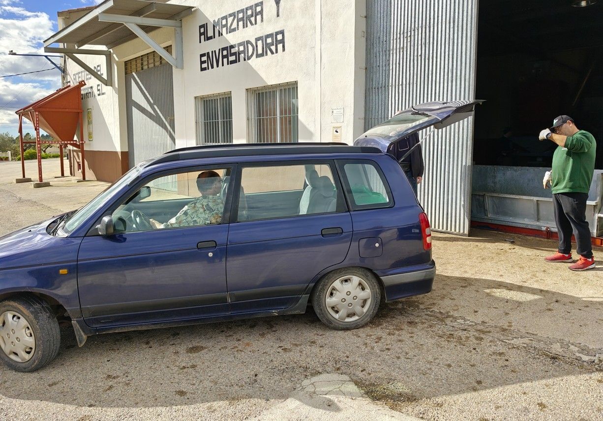 Een blauwe auto staat geparkeerd voor een gebouw. ​​Mensen laden spullen in de open kofferbak. Op het gebouw staat een bord met 