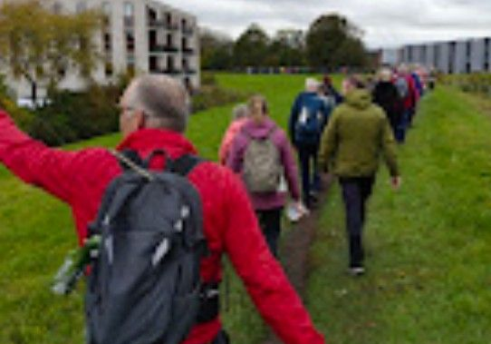 Een groep mensen loopt over een pad, aangevoerd door een persoon in een rood pak, met gebouwen en gras op de achtergrond.