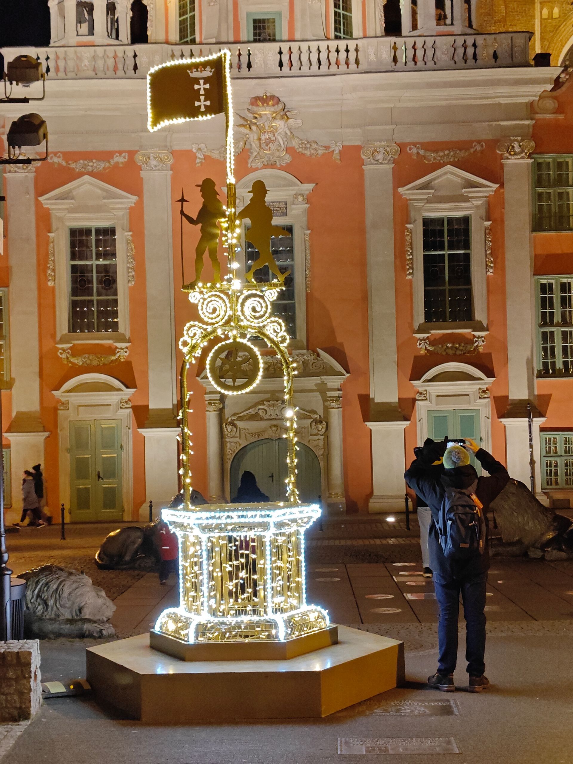 Verlicht, putvormig beeldhouwwerk met figuren en een vlag voor een gebouw; iemand maakt een foto.