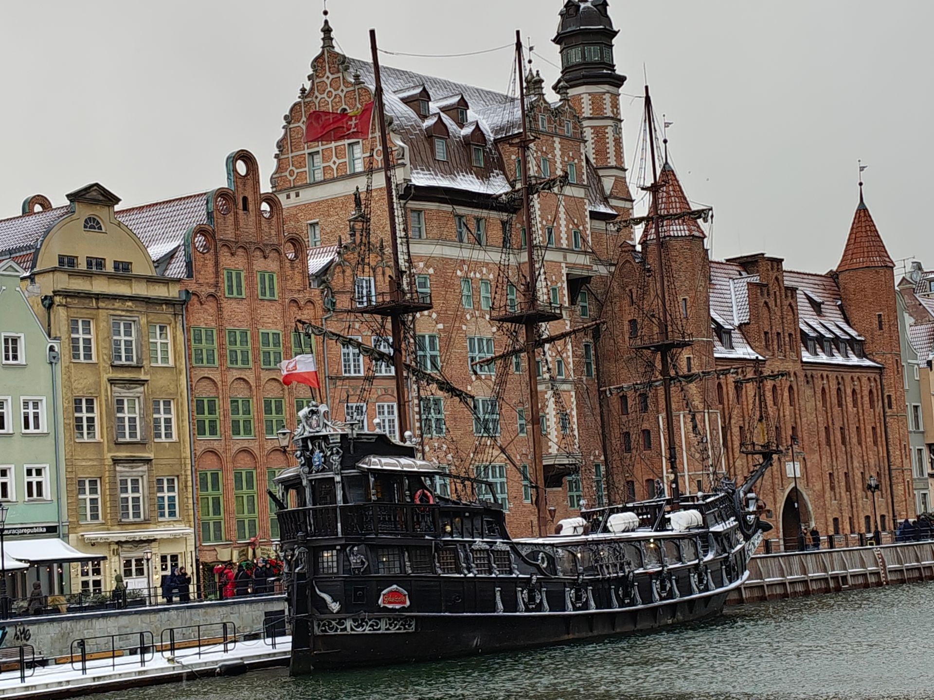 Een zwart zeilschip ligt aangemeerd in een kanaal, met kleurrijke gebouwen op de achtergrond onder een bewolkte hemel.