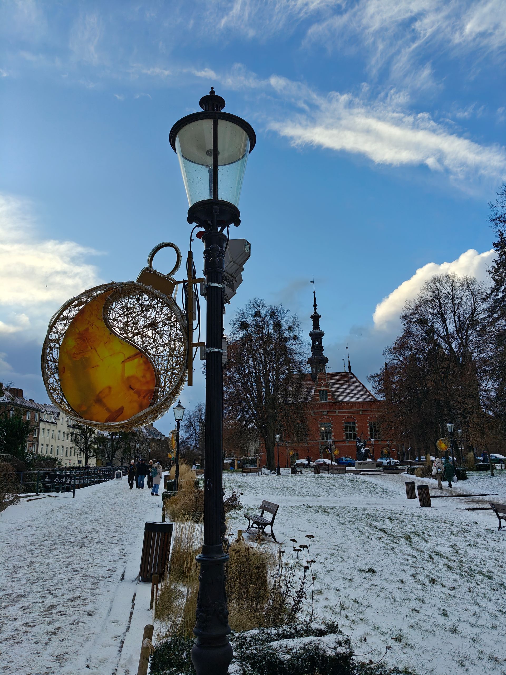 Een besneeuwd park met een lantaarnpaal en een kerstversiering. Op de achtergrond een rood gebouw en een blauwe lucht.