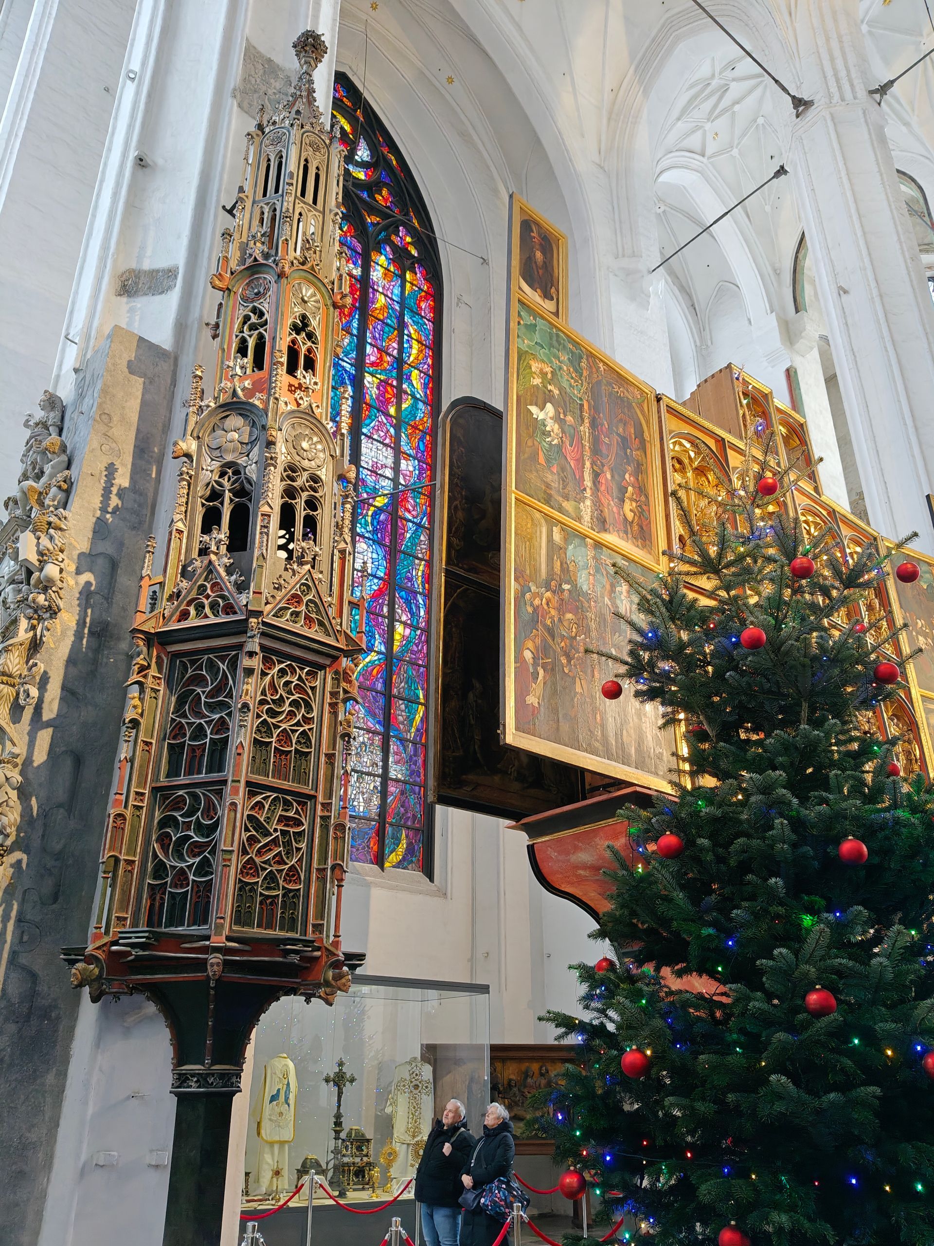 In een kerk staat een kerstboom bij het altaar, versierd met glas-in-loodramen en houtsnijwerk.