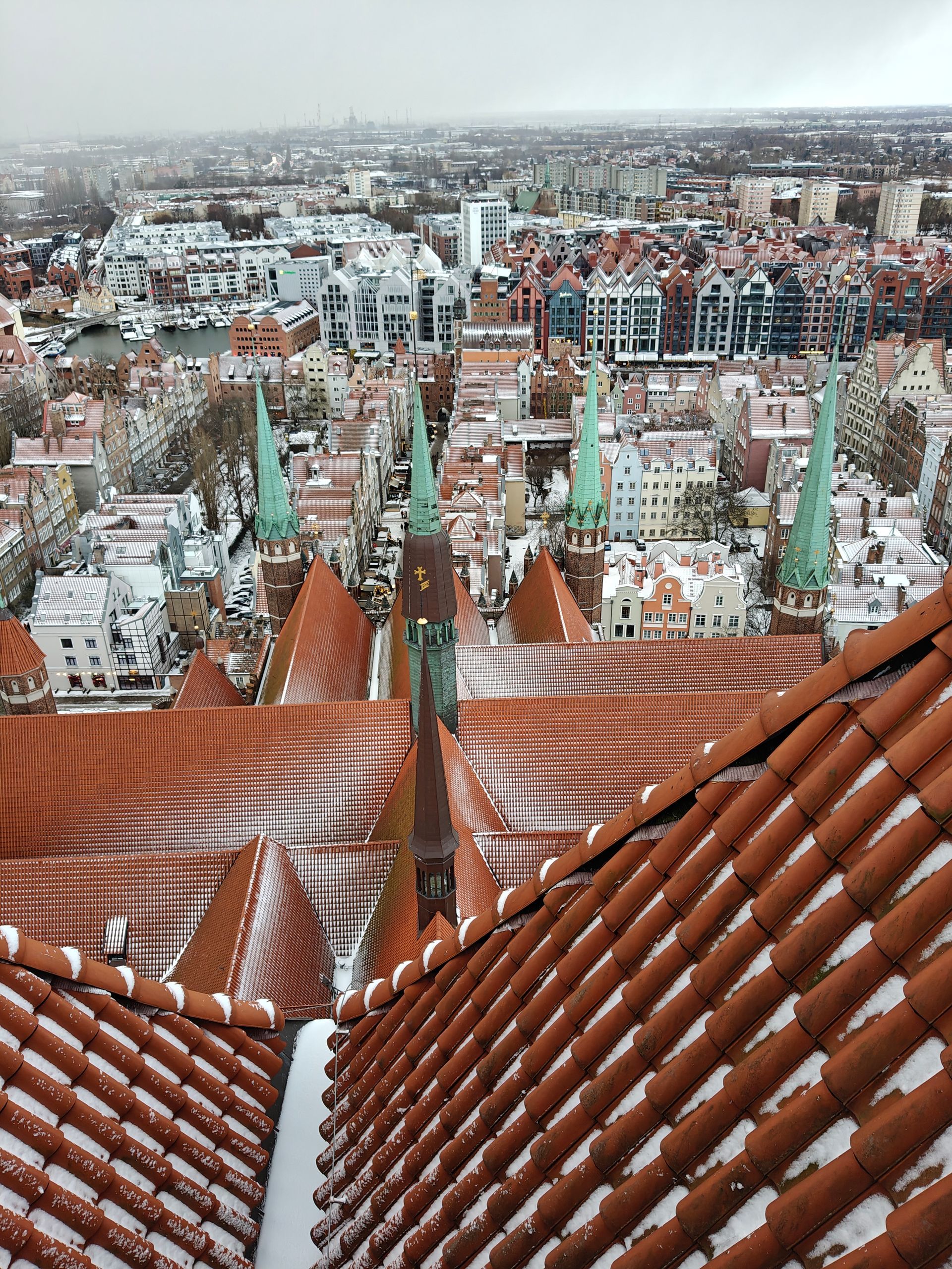 Uitzicht vanaf het dak op een met sneeuw bedekte stad met oranje pannendaken en groene gebouwen met spitsen.