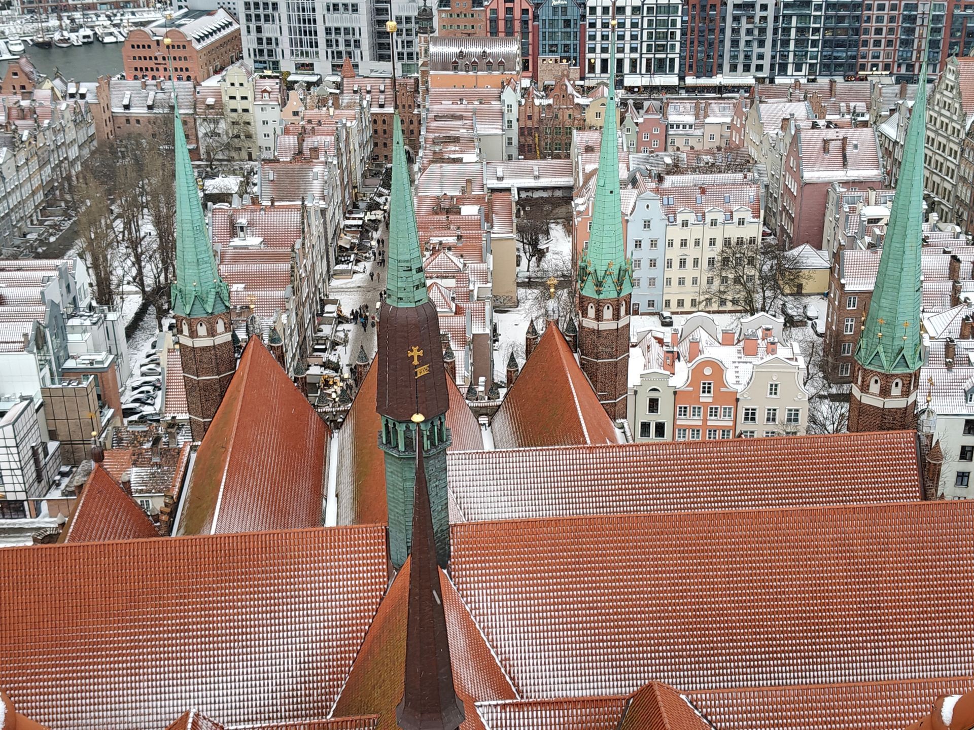 Een hoog perspectief van een stad met rode pannendaken, groene torens en sneeuw. De gebouwen staan ​​dicht op elkaar.