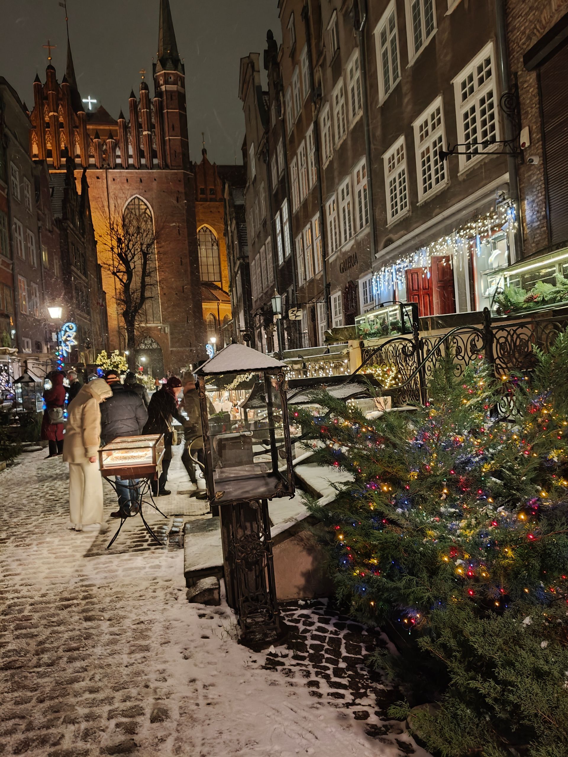 Een besneeuwde straat met kinderkopjes, een kerstboom, versierde gebouwen en een kerk op de achtergrond, 's nachts.