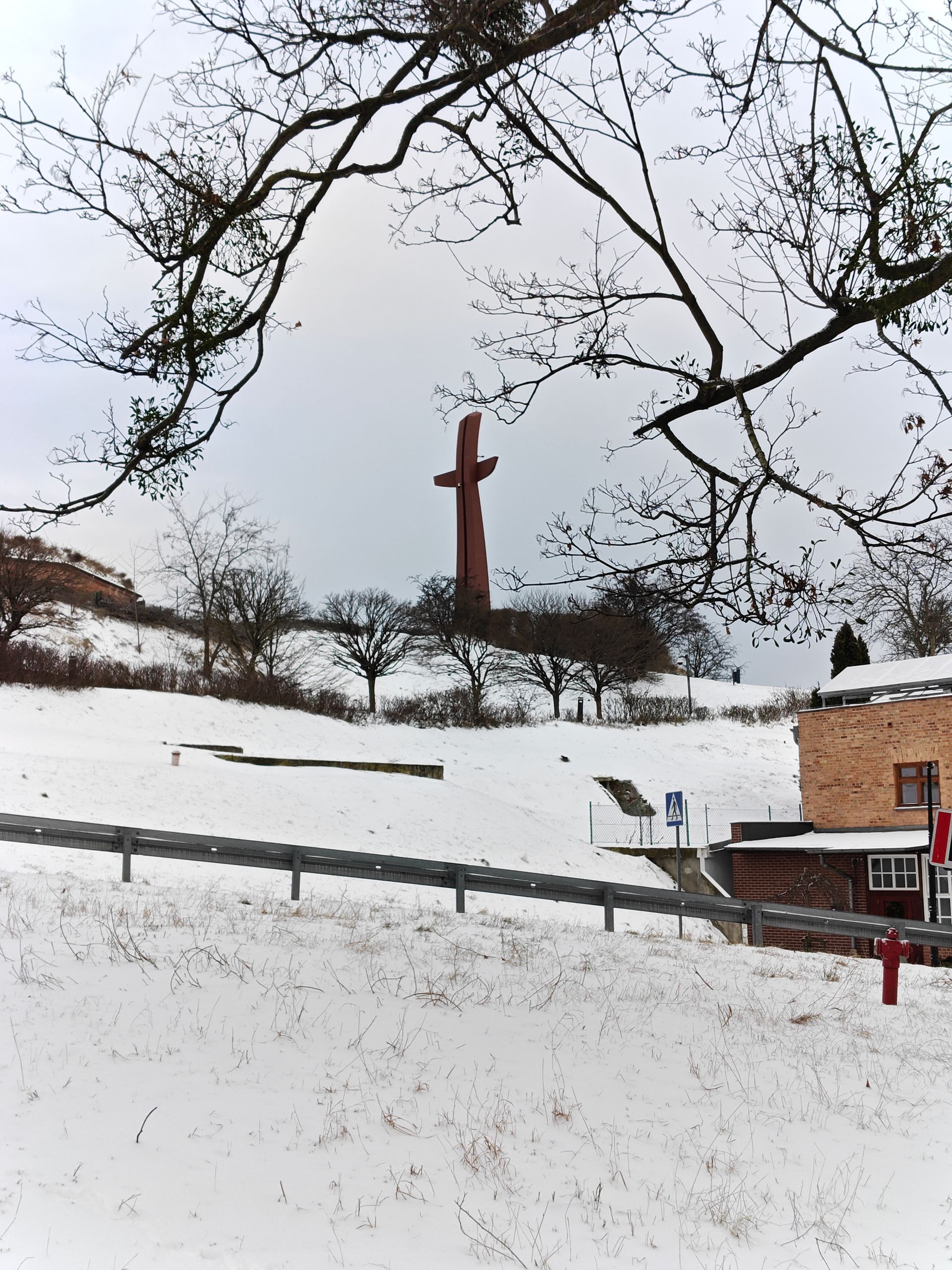 Een besneeuwd landschap met in de verte een hoog, roodbruin beeldhouwwerk. Kale bomen en een gebouw zijn zichtbaar.