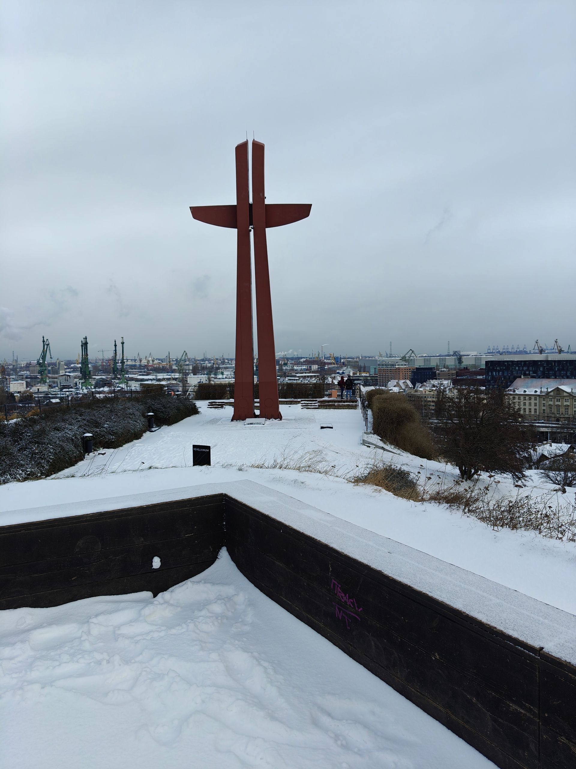 Een besneeuwd landschap met een groot, bruin kruisbeeld op een heuveltop. De skyline van de stad is in de verte zichtbaar.